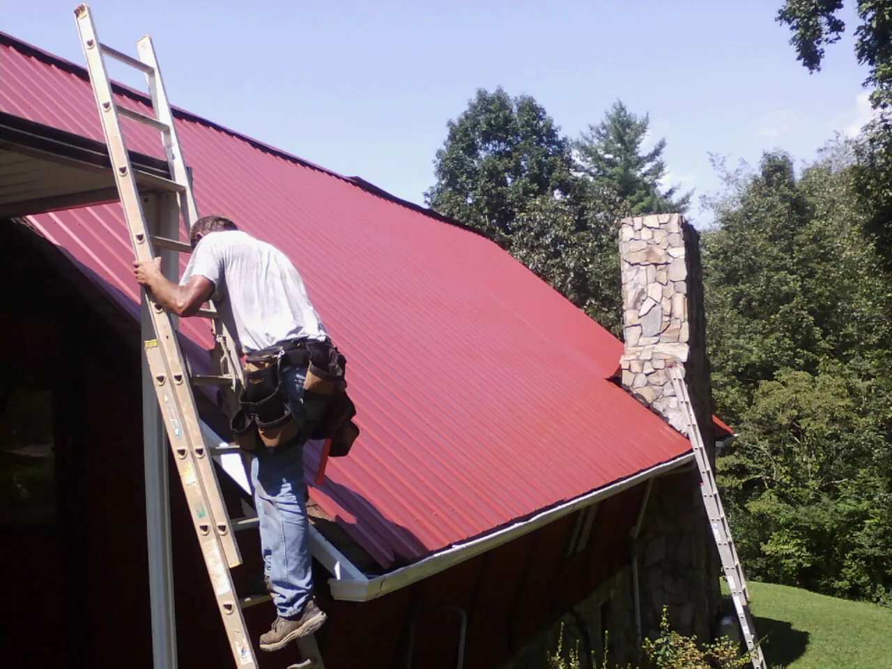 Roofer on a ladder beside a red metal roof and stone chimney — Soffit & Fascia Repair by Roofing Brothers in Montague